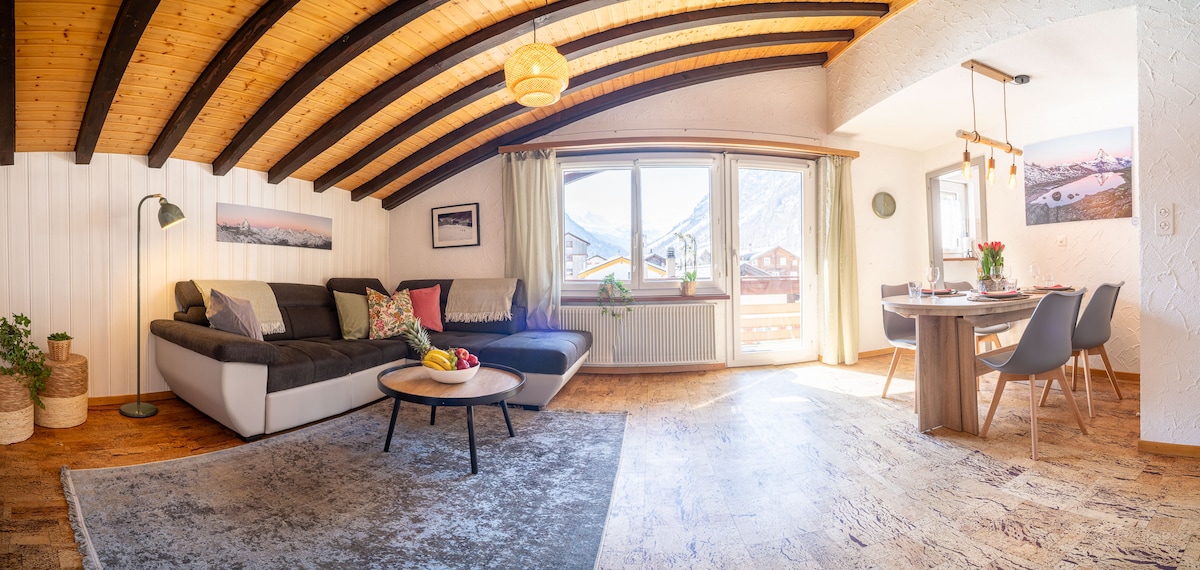 A welcoming living area features a comfortable sectional sofa adorned with decorative pillows, complemented by a round coffee table. Sunlight filters through large windows, illuminating the space. A dining table with modern chairs and fresh flowers adds a touch of warmth, enhancing the inviting ambiance.