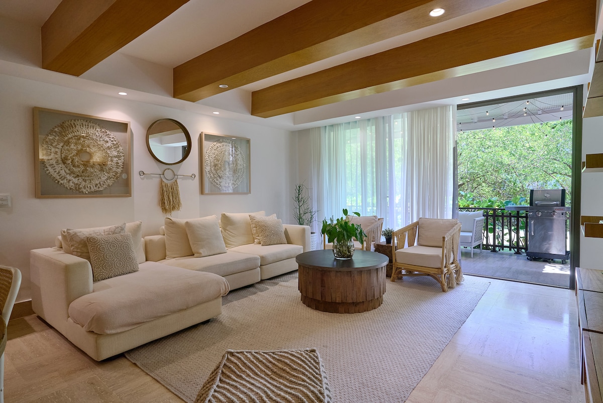 A spacious living room features a light-colored sectional sofa and a round wooden coffee table. Natural light flows through large windows adorned with sheer curtains, illuminating the open space. A cozy seating area is visible, inviting relaxation and conversation.