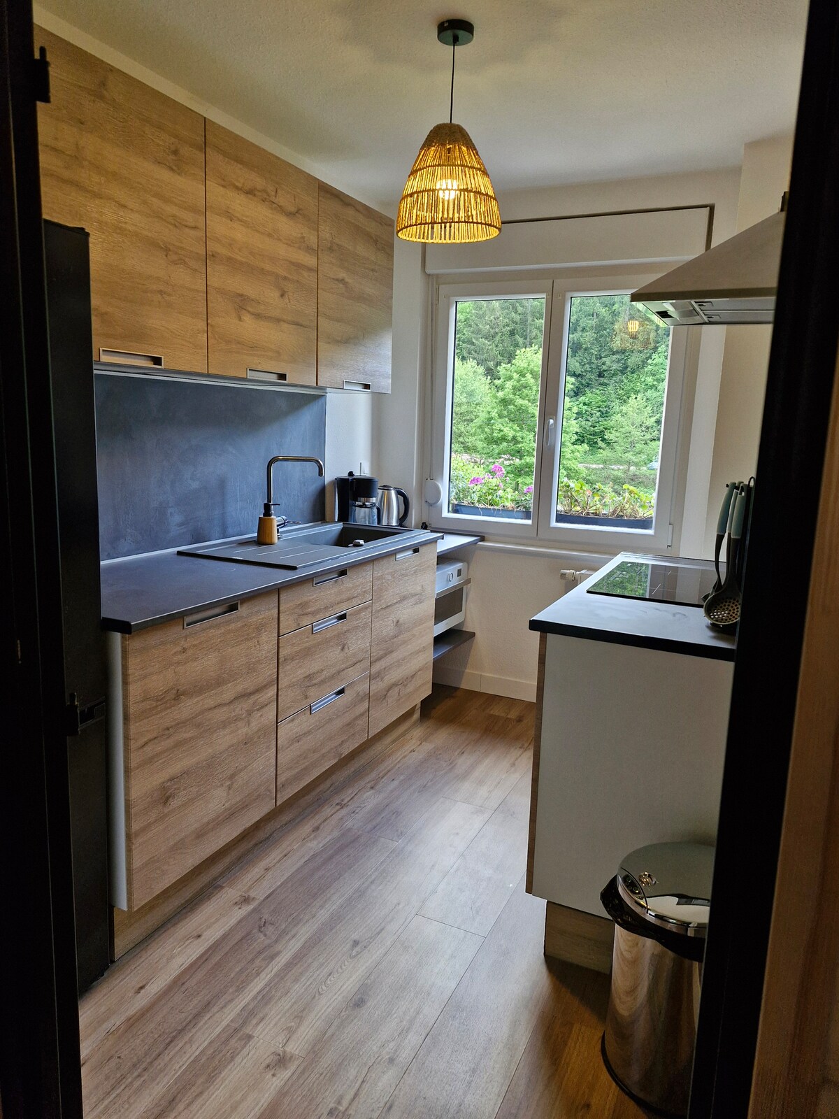 A modern kitchen is shown, featuring wooden cabinetry and a black countertop. A window allows natural light to enter, revealing lush greenery outside. Appliances, including a kettle and a coffee maker, are neatly arranged, while a metal trash can is positioned in the corner.