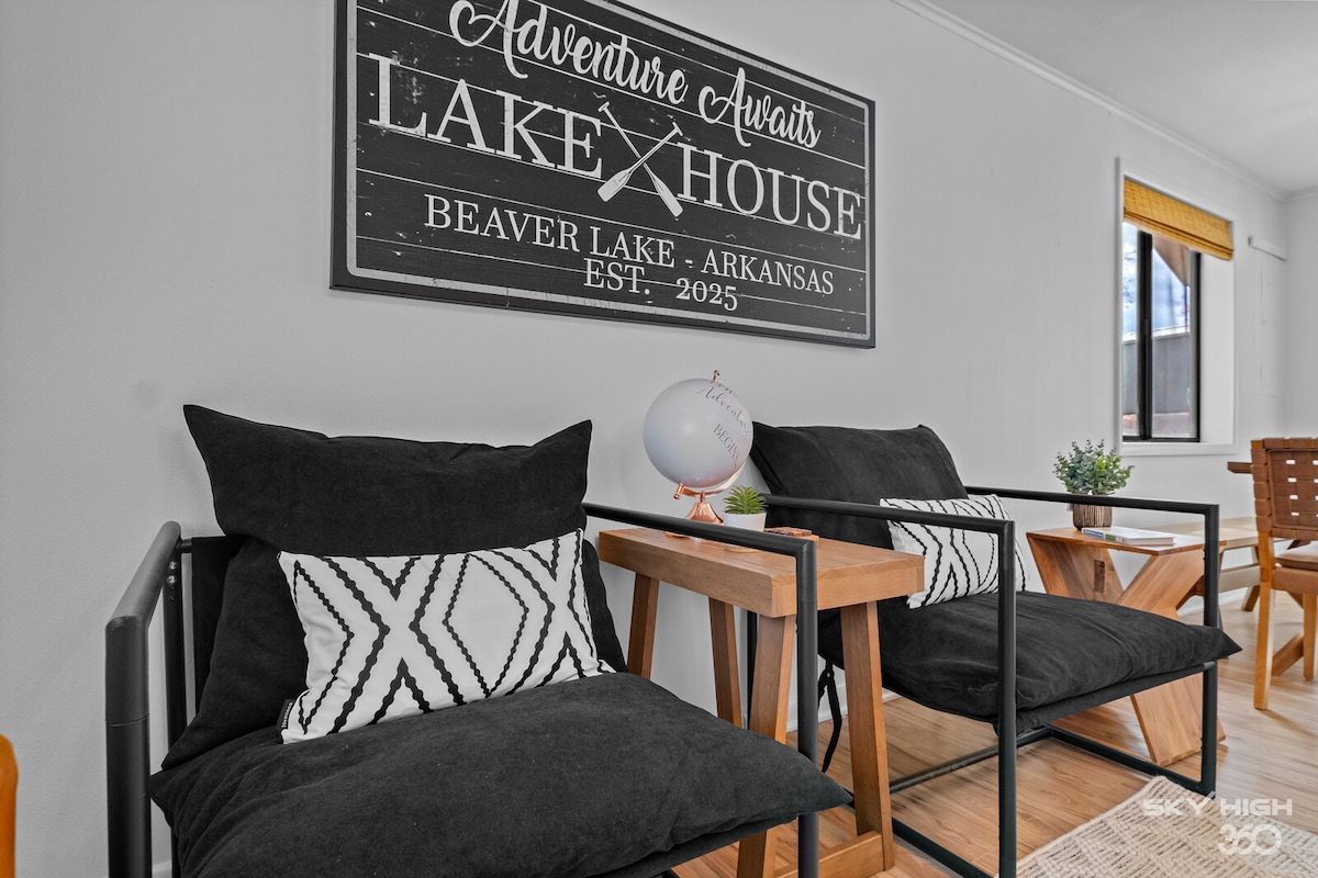 Two modern chairs with black cushions and wooden side tables are placed near a wall featuring a decorative sign that reads 'Adventure Awaits Lake House Beaver Lake, Arkansas.' Natural light enters through a nearby window, illuminating the inviting seating area.