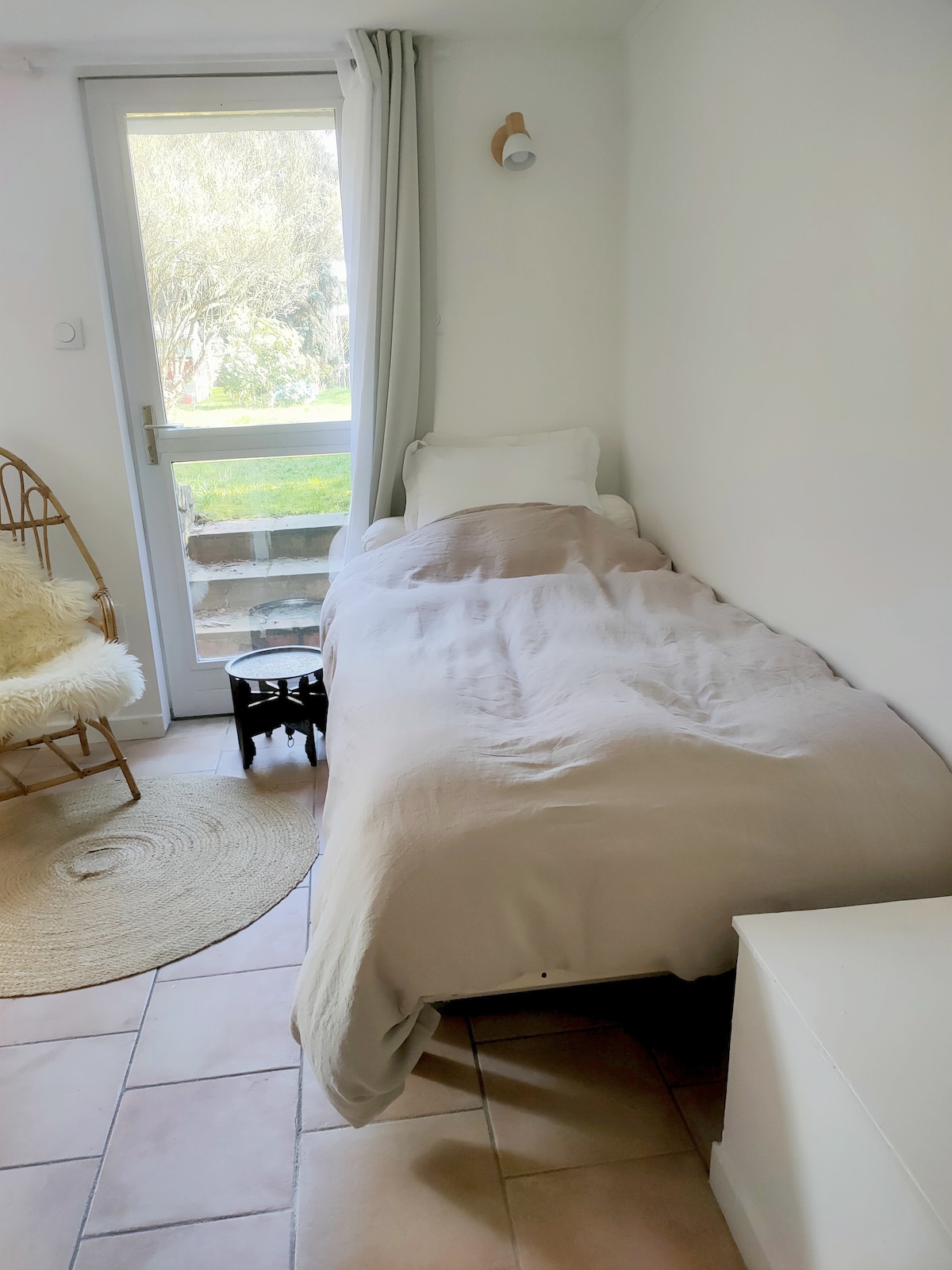 A compact sleeping area features a single bed with a light quilt, positioned alongside a stylish side table. A wicker chair and a round rug add warmth to the space, while a glass door provides a view of the garden outside.
