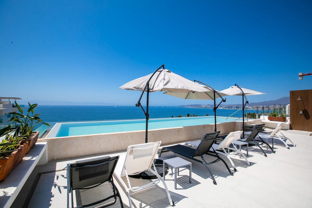 An outdoor lounge area is presented, featuring white umbrellas and black and white lounge chairs arranged around an infinity pool. The view showcases the vibrant blue sea and clear sky, with the coastline visible in the distance.