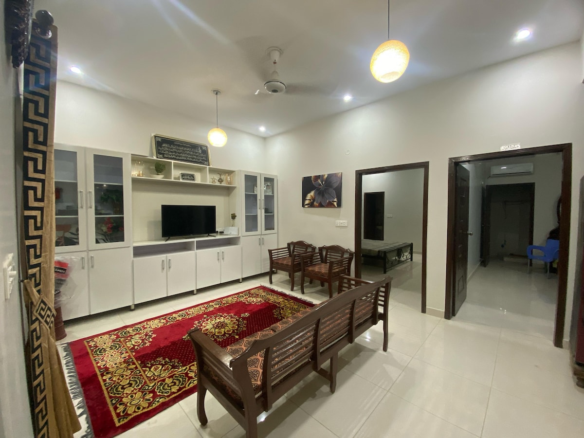 A spacious living area is furnished with a set of wooden chairs arranged around a central area rug. White cabinets house a television, while modern lighting fixtures provide ample illumination. Neutral walls and tiled flooring contribute to a bright and open atmosphere, complemented by an entrance visible to the side.