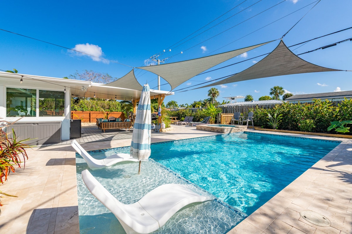 A private outdoor space features a shimmering pool, complemented by two lounge chairs partially submerged in the water. Patio shades provide relief from the sun, while tropical greenery surrounds the area, offering a tranquil atmosphere for relaxation. Comfortable seating can be seen nearby.