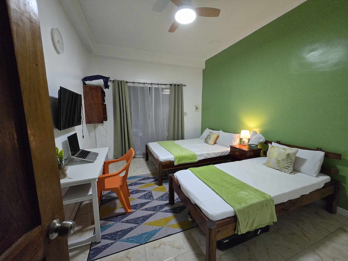 חדר שינה מואר עם שתי מיטות נפרדות עם מצעים לבנים ושטיחי מסדרון ירוקים, על רקע קיר ירוק בולט. החדר כולל שולחן לילה עם מנורה, טלוויזיה המורכבת על הקיר, שולחן עבודה עם כיסא כתום ושטיח גיאומטרי צבעוני.