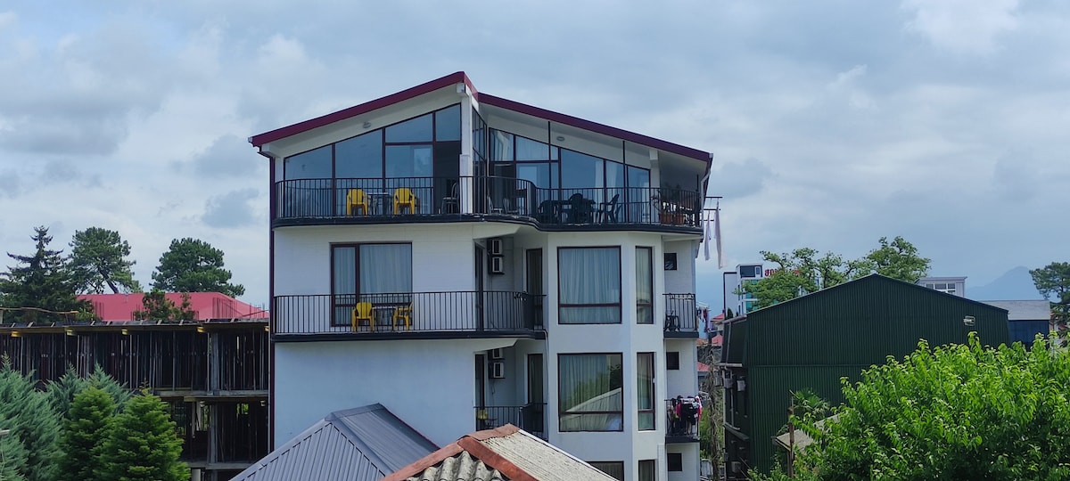A three-story building features large glass windows and a sloped roof. Balconies are visible on each floor, offering outdoor seating. Lush greenery surrounds the structure, contributing to a serene environment. Nearby buildings and a clear sky with scattered clouds are also present.