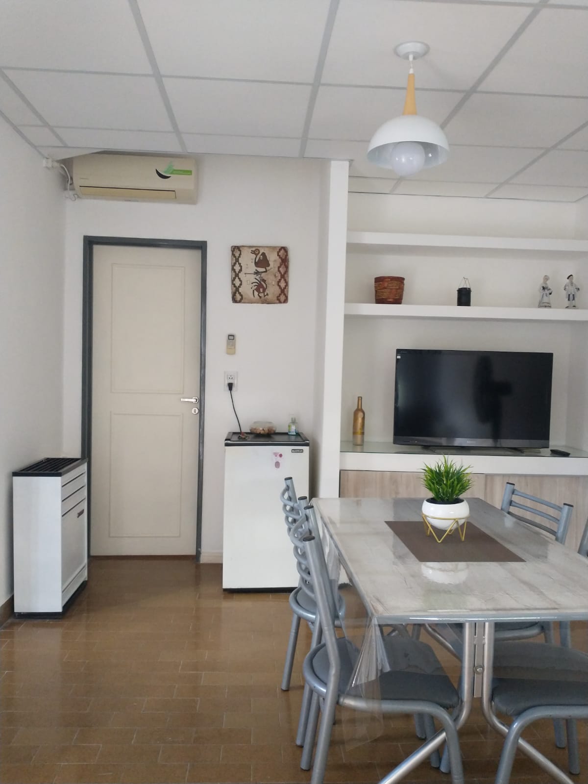 A dining area is presented with a rectangular table surrounded by six metal chairs. A small refrigerator is positioned next to the table. A television is mounted on a wall-mounted shelf, featuring decorative items. An air conditioning unit is visible above the entry door.