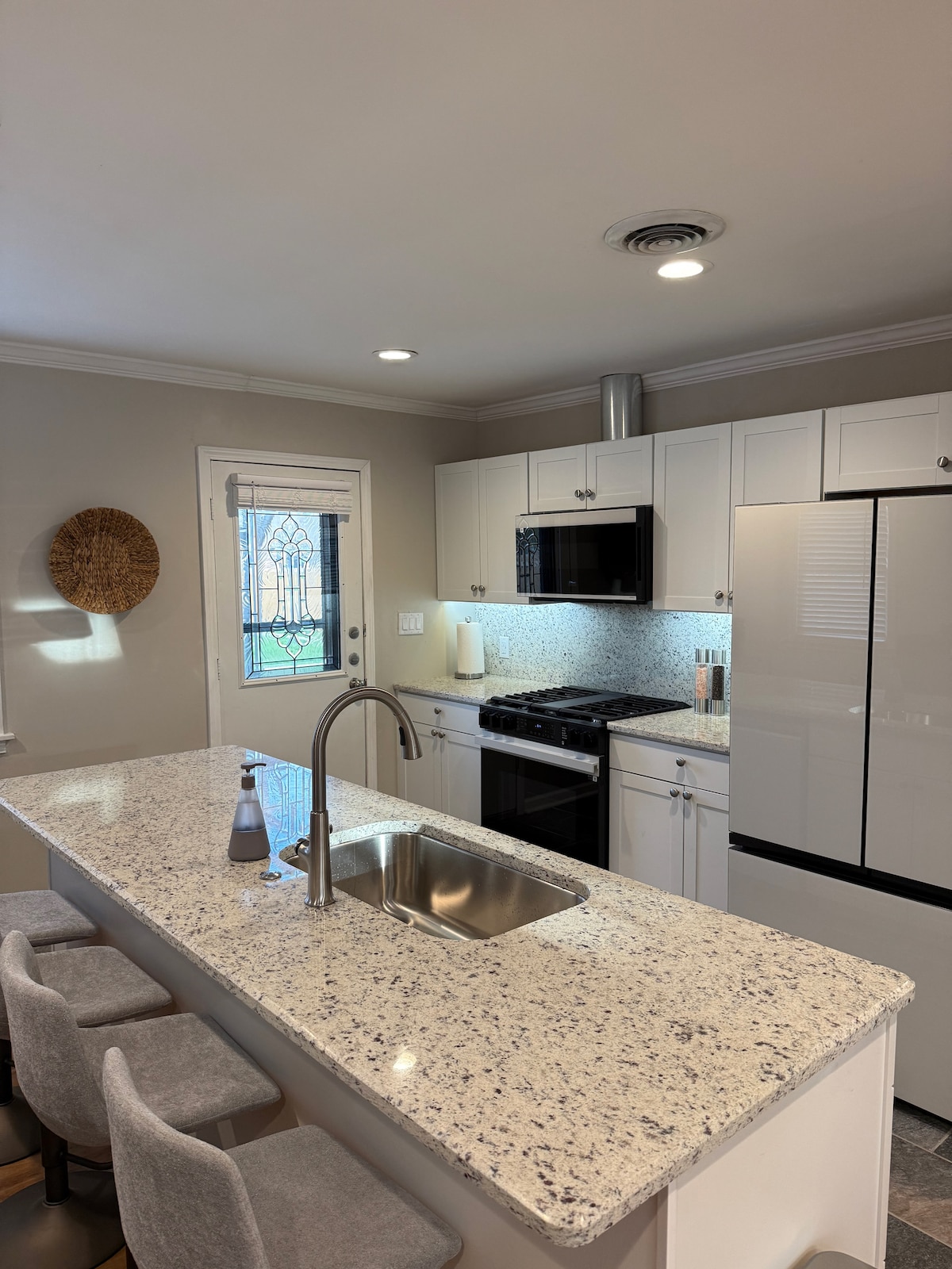 A modern kitchen features granite countertops and stainless steel appliances. A central island with seating is complemented by a sleek sink. Natural light comes through a nearby window, while a woven wall decor piece adds a subtle touch to the space.