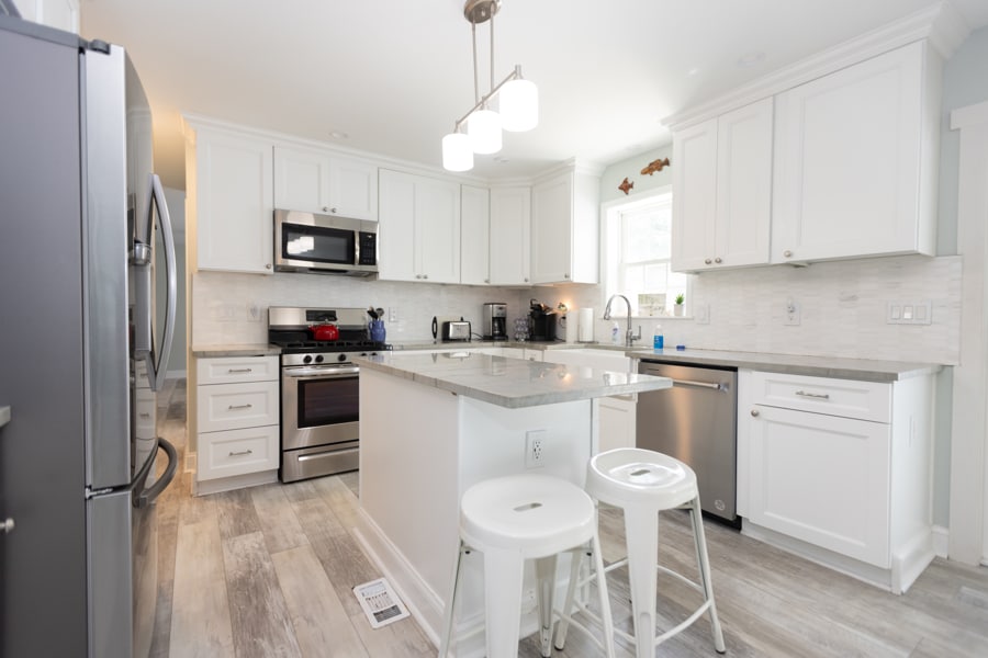 A modern kitchen features sleek white cabinetry and a spacious island with seating. Stainless steel appliances are complemented by gray countertops and light-colored flooring. Natural light fills the room through a window, enhancing the welcoming ambiance.
