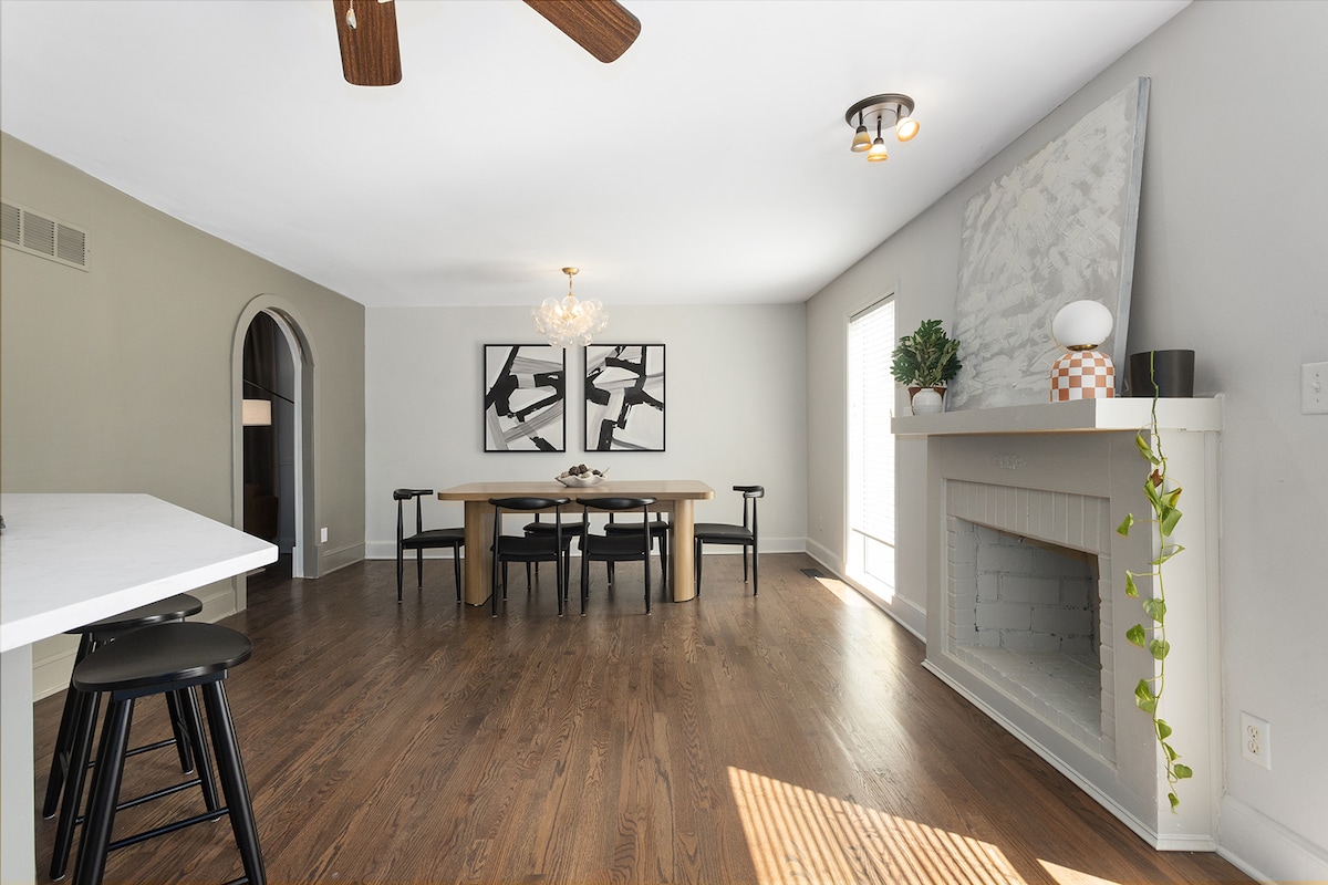 The dining area features a long rectangular table surrounded by black chairs, designed for group gatherings. A fireplace is situated on one side, while large windows allow natural light to fill the room. A modern chandelier provides additional illumination, and artwork adorns the walls.