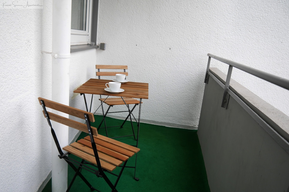 A small balcony area features a wooden table set with two matching folding chairs. Each chair has a cup placed on the table. The floor is covered with green outdoor carpeting, and the walls are painted white, creating a clean and simple space.