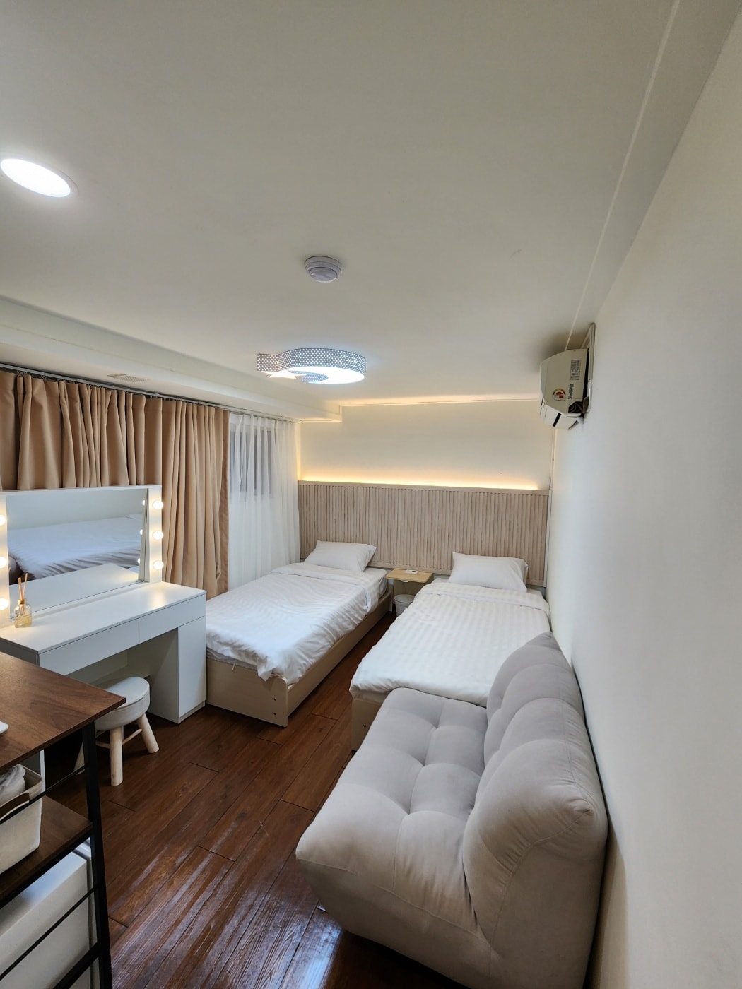 A well-lit bedroom features two single beds with light bedding. A cozy sofa sits in the corner, complemented by a stylish vanity area adorned with lights. Large windows are framed by soft curtains, allowing natural light to brighten the serene space.