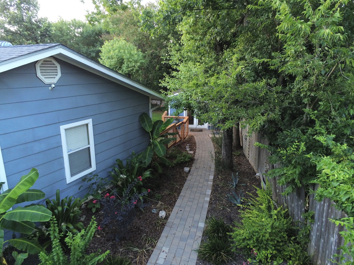 A paved pathway winds through a lush garden, flanked by vibrant green plants and colorful flowers. The guest house, painted in a soft blue hue, is visible at the side, along with a wooden deck beyond the trees, inviting a serene outdoor experience.
