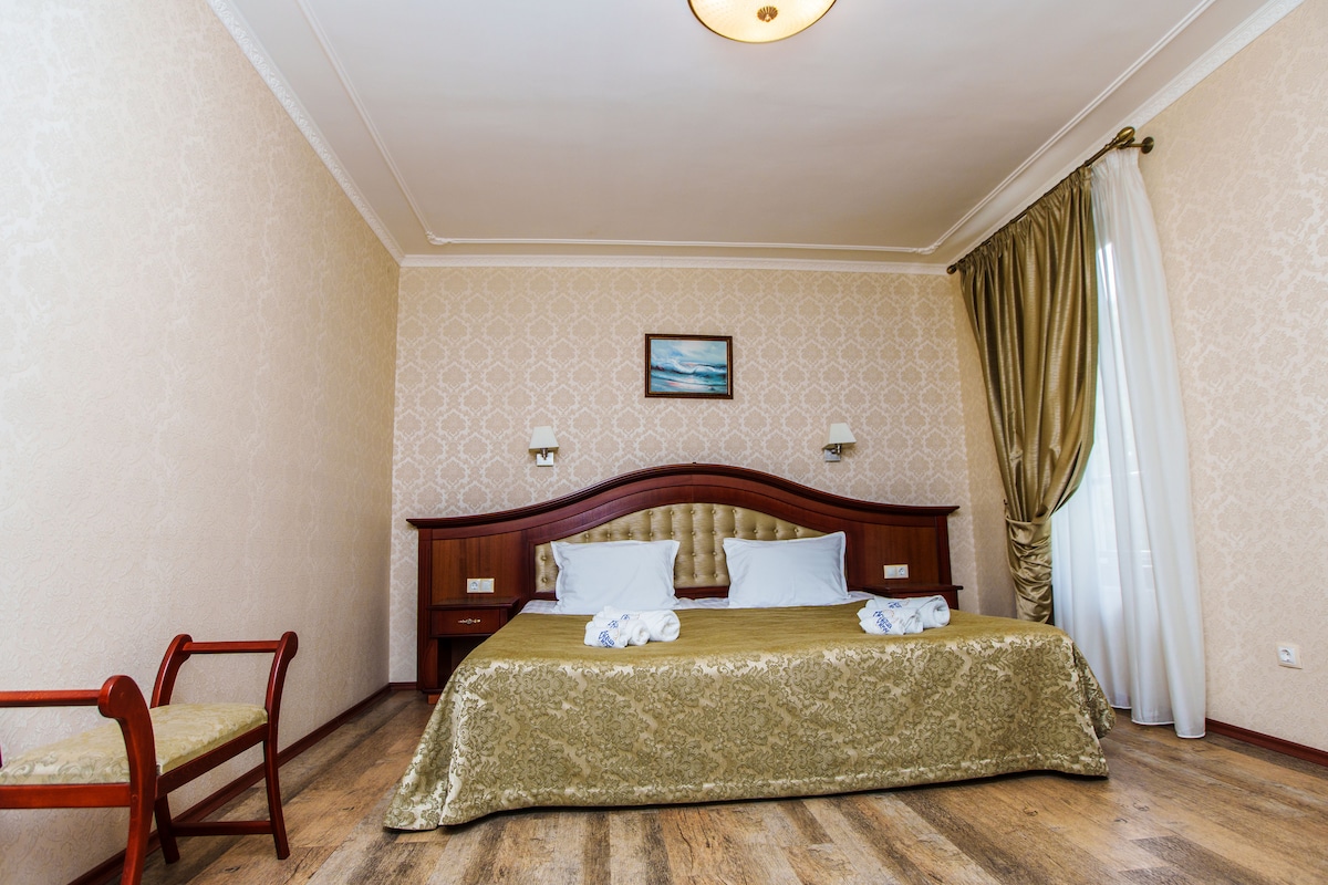 The hotel room features a spacious bed with soft linens and complementary pillows. A small wooden chair is placed beside the bed. Natural light filters through sheer curtains, illuminating the warm tones of the décor. A piece of artwork hangs above the headboard.