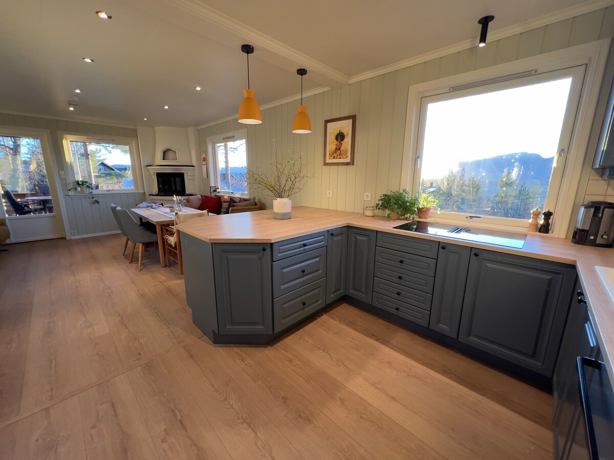 A modern kitchen is featured with a spacious countertop and grey cabinetry. Pendant lights illuminate the area, and a large window offers a view of the outdoors. The dining area is visible, showcasing a table with chairs and a cozy seating corner.