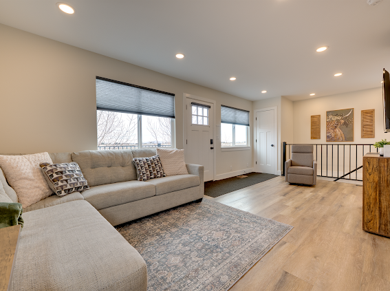 A spacious living area is filled with soft natural light from large windows. A light grey sectional sofa is paired with decorative pillows, while a plush area rug lies beneath. A comfortable armchair and a wooden TV stand complete the inviting atmosphere.