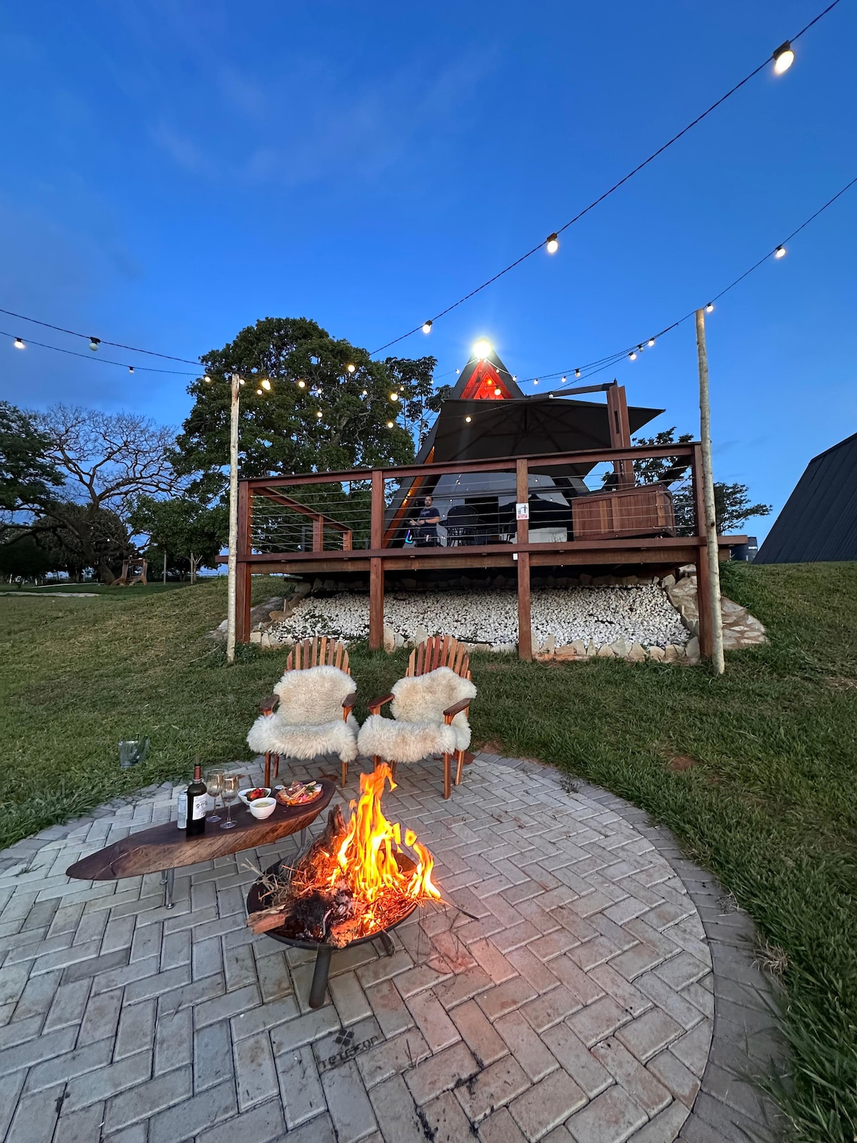 An outdoor fire pit is surrounded by two wooden chairs, each draped with a cozy throw. Soft lighting from string lights above enhances the setting, while a grassy area and a patio structure create a welcoming atmosphere for relaxation and gatherings.