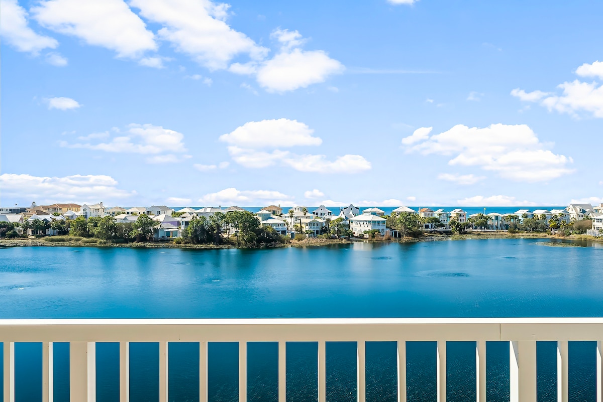 A wide-angle view displays serene waters and charming homes along the shoreline. Blue skies dotted with fluffy white clouds create a peaceful backdrop. The balcony railing is visible in the foreground, enhancing the sense of outdoor space and tranquility.