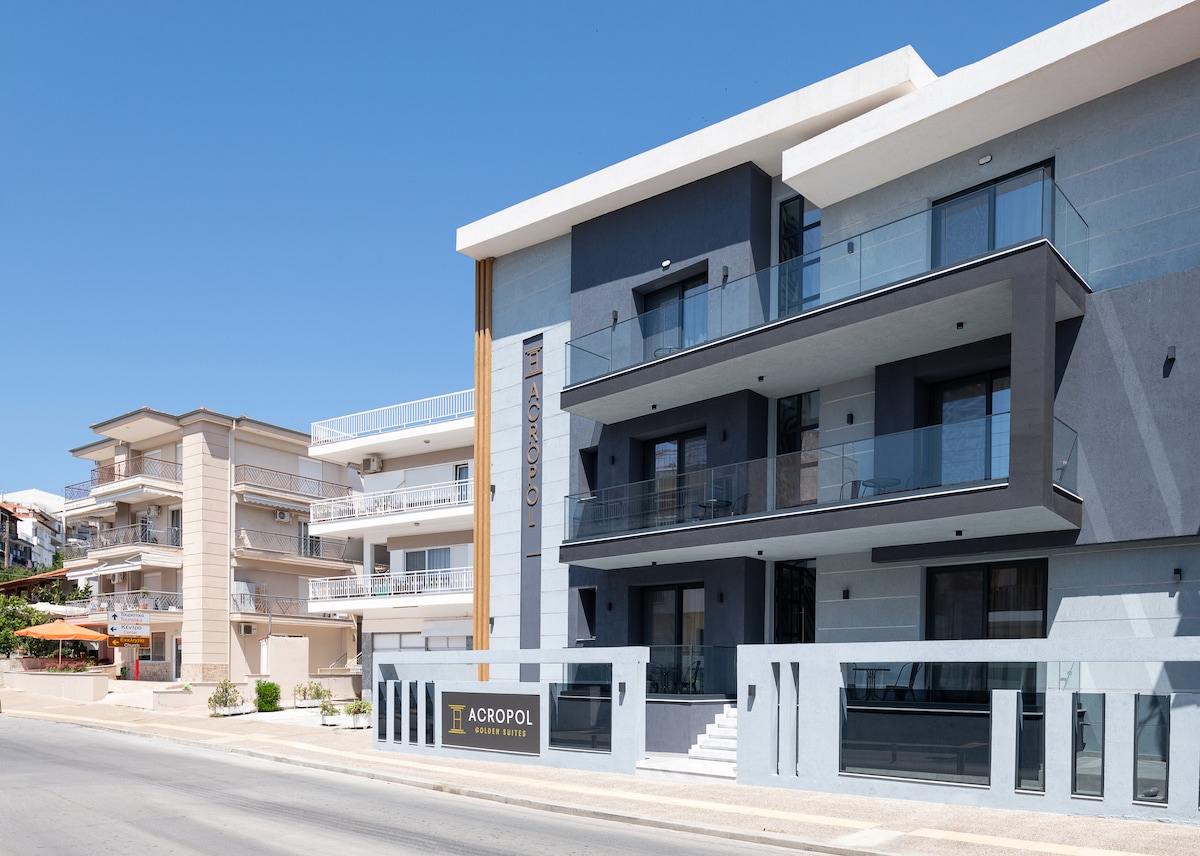 The exterior of Acropol Golden Suites is displayed, featuring a modern three-story building with a sleek facade. Balconies with glass railings are visible, and the entrance is marked by the property's name on a stylish sign. Surrounding structures are also seen along the street.