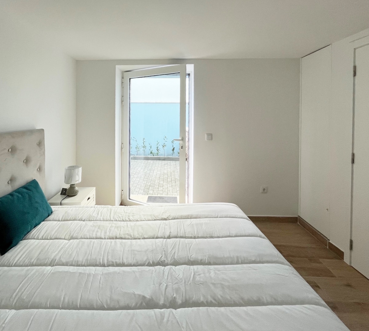 A bright, inviting bedroom features a spacious bed with crisp white bedding and a teal accent pillow. Large glass doors open to a private terrace, allowing natural light to fill the room. A bedside lamp and minimalistic furniture contribute to a clean, modern aesthetic.