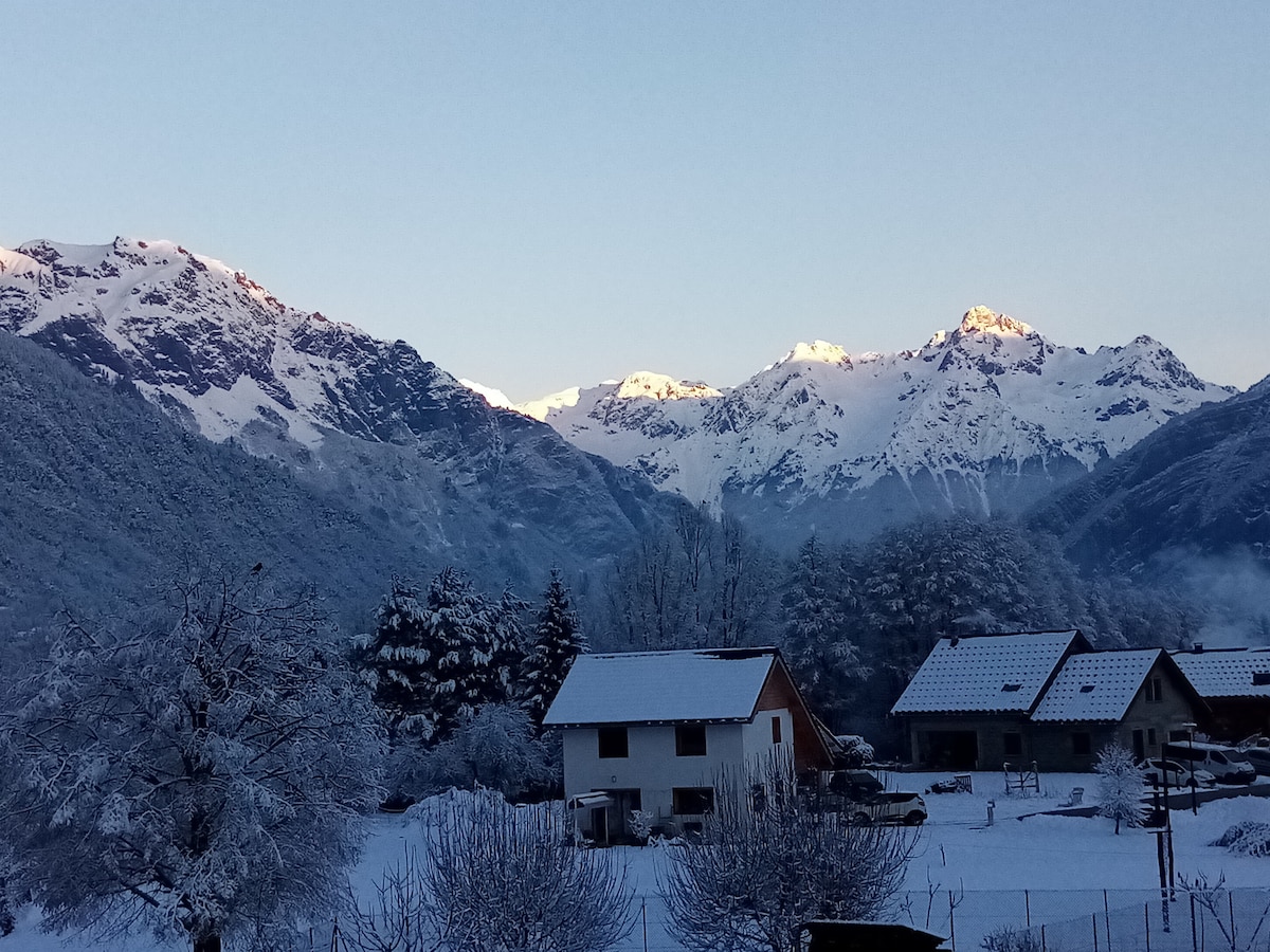 A snow-covered landscape features tranquil mountains in the background, with peaks reflecting soft sunlight. A quaint white house is visible in the foreground, surrounded by frost-coated trees and a peaceful winter atmosphere.
