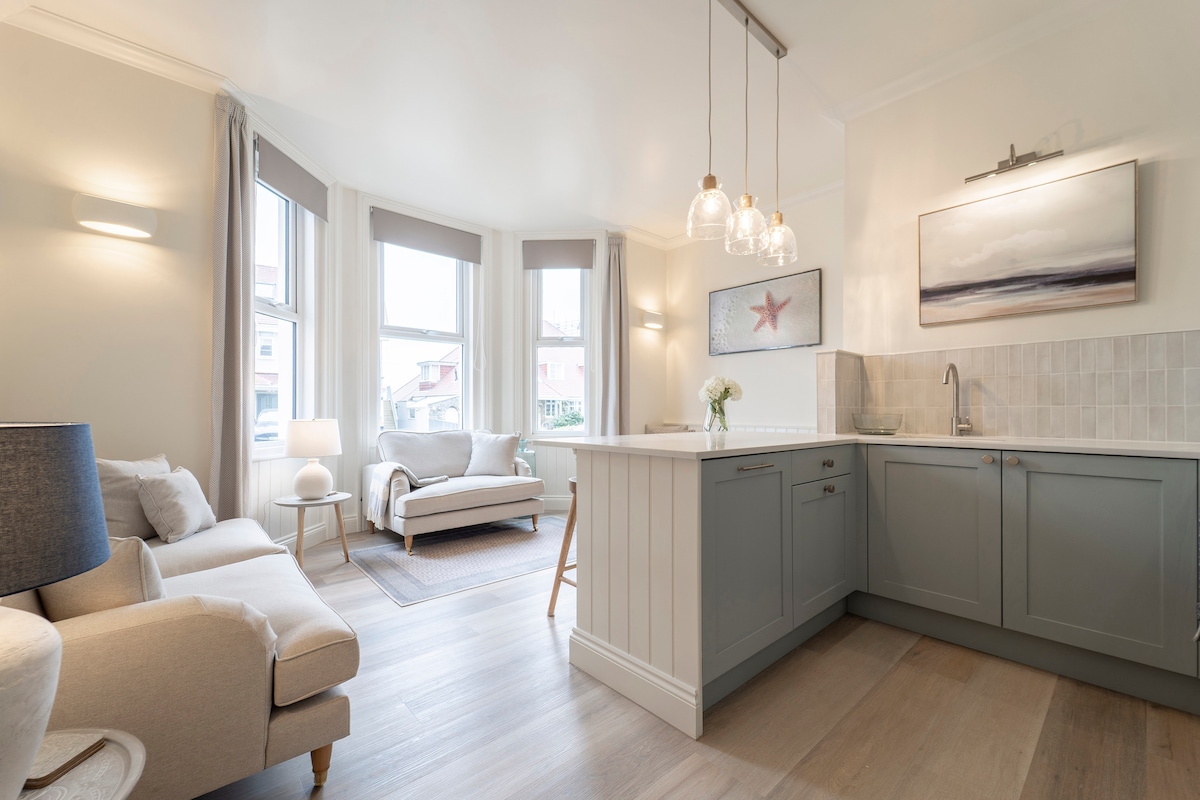 A light-filled open-plan living space features a cozy sofa and a stylish kitchen island with contemporary pendant lighting. The walls are painted in soft tones, while large windows allow natural light to illuminate the room. Decorative elements like a flower in a vase and coastal artwork add charm.