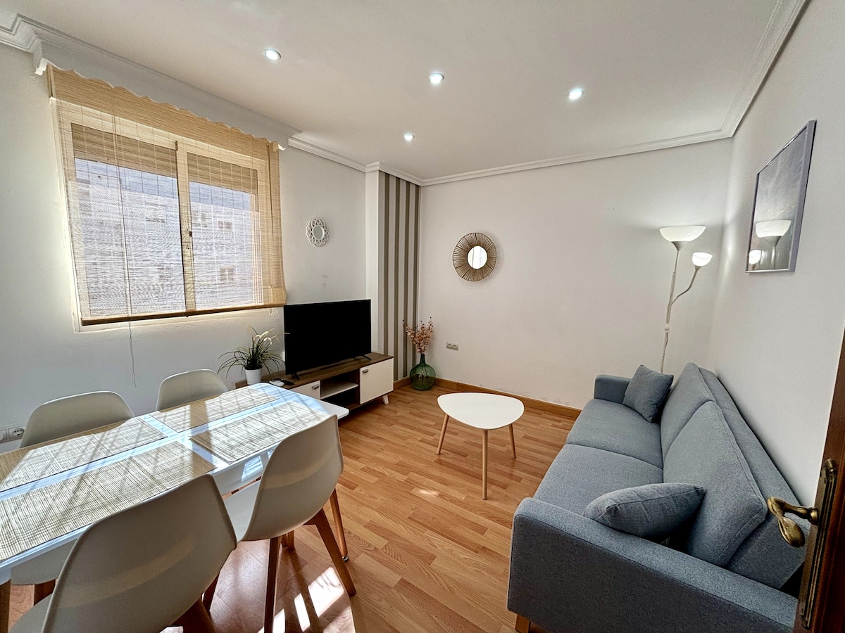 A living area features a comfortable gray sofa and a small round table, surrounded by four light-colored chairs. A television is positioned on a low media console. Bright light filters through large windows covered with light fabric, illuminating the wooden floor.