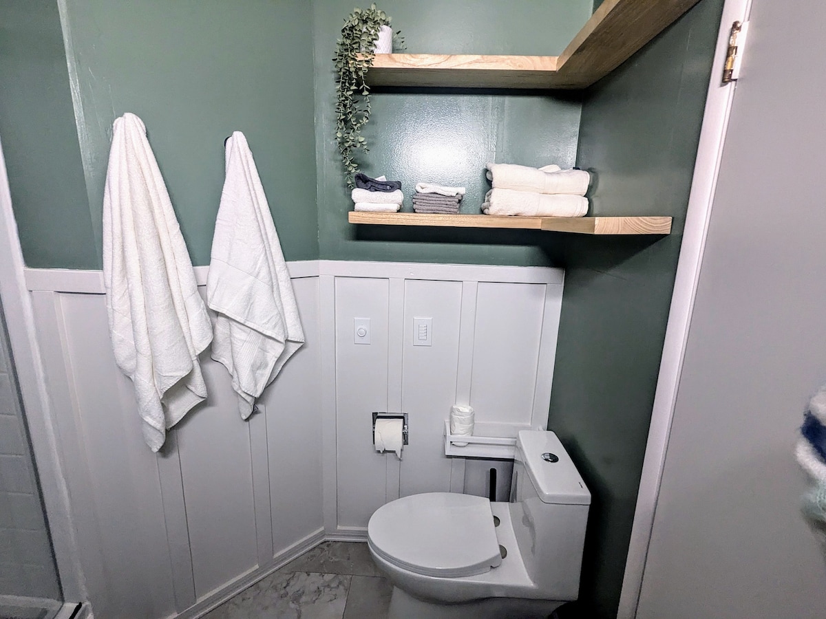 A clean bathroom features light green walls and a neutral tile floor. Two neatly arranged towels are hung on hooks. Shelves hold stacked towels and toiletries, while a modern toilet is positioned beside a minimalistic wall design.