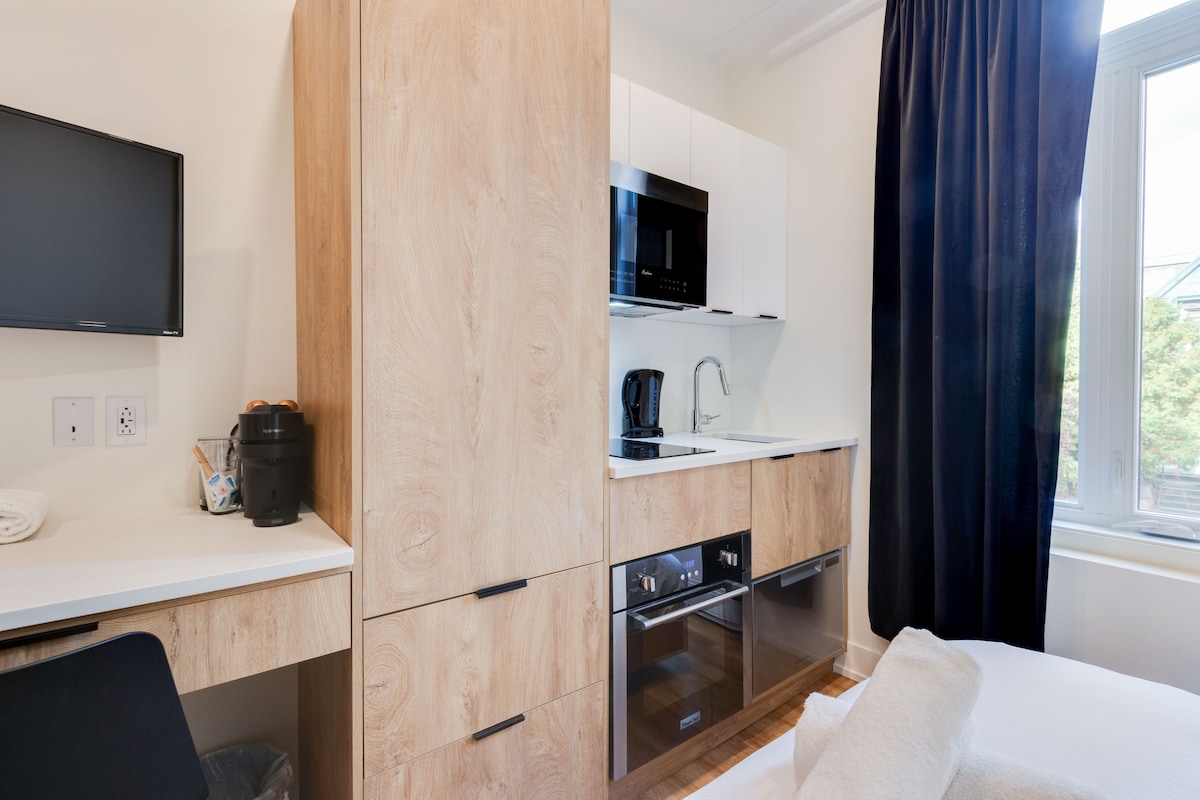 A modern kitchenette features a microwave, oven, and coffee maker, integrated into sleek cabinetry. A small desk area is visible, along with a flat-screen TV mounted on the wall. A large window with dark curtains allows natural light to brighten the space.