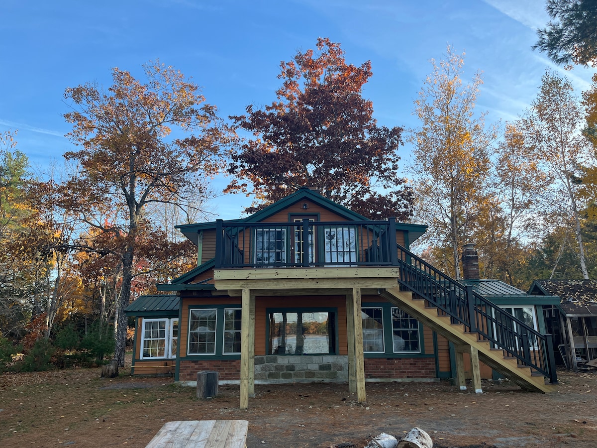 The cabin is seen from the front, showcasing its two-story design with a staircase leading to the upper level. Surrounding trees exhibit vibrant autumn foliage, complementing the rustic exterior. A wooden deck above extends over the base level, inviting outdoor relaxation.