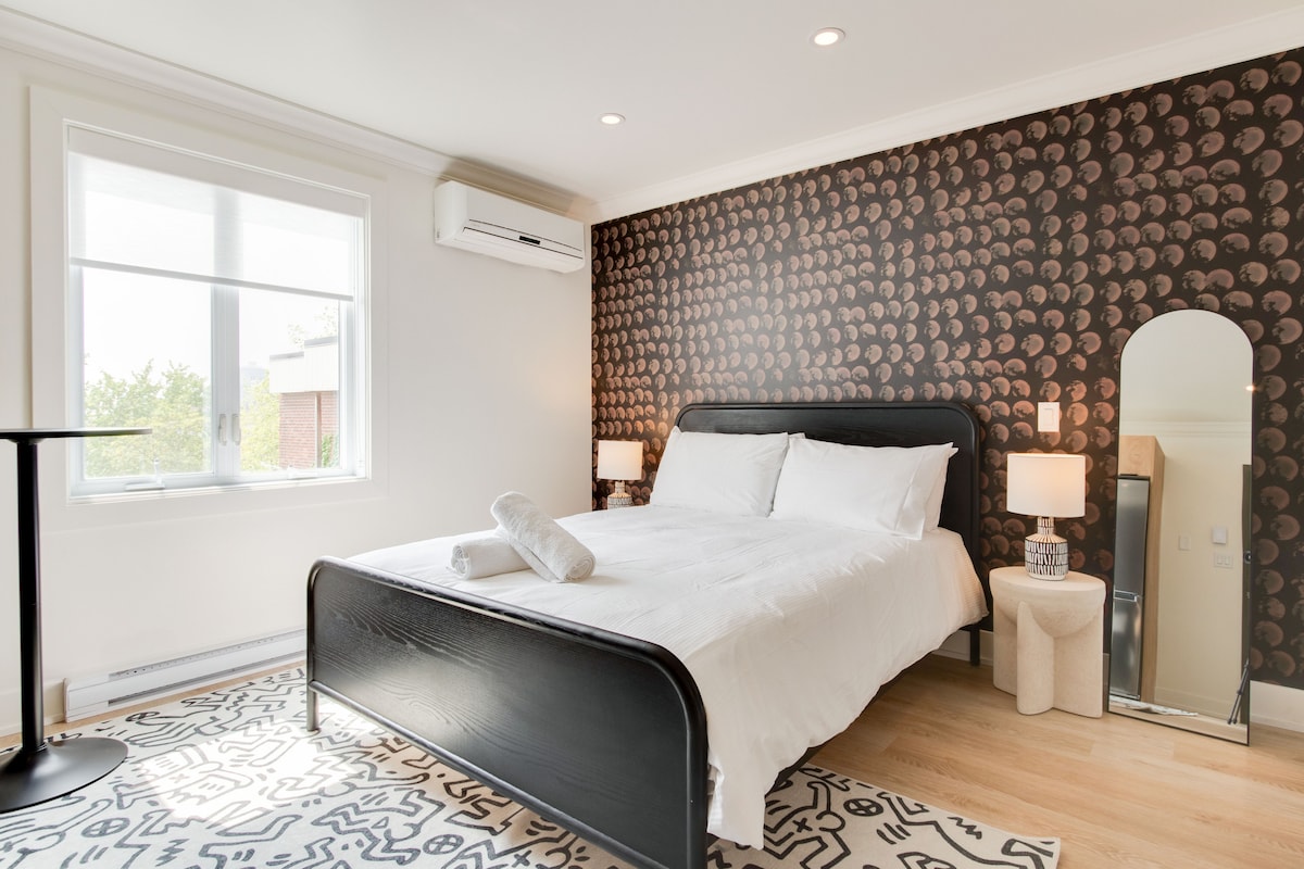 A modern bedroom features a plush queen bed dressed in crisp white linens. An accent wall with a unique design contrasts with the light wood flooring. Natural light fills the space through a large window, while a mirror and bedside lamps add functionality and style.