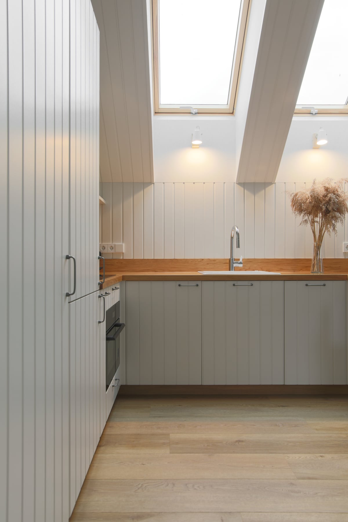 A modern kitchen features light wooden accents and vertical panelled cabinetry. Natural light is provided by skylights above, illuminating the space. A sleek countertop is complemented by a sink and a simple arrangement of dried plants in a vase, enhancing the kitchen's contemporary design.