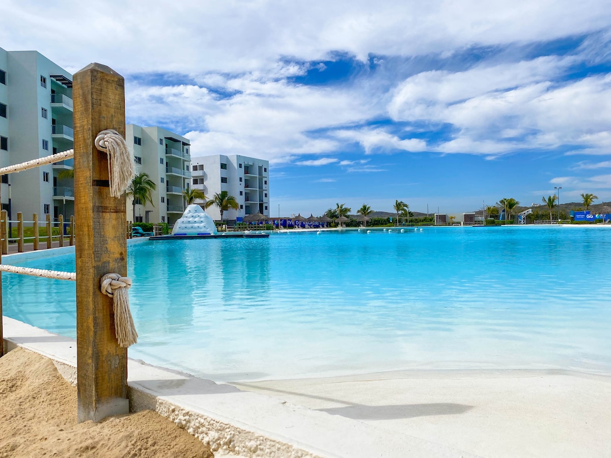 A serene artificial lagoon is depicted, with clear turquoise water reflecting the sky. Adjacent to the lagoon, modern buildings are visible in the background. A section of sandy shore frames the water's edge, creating a tranquil setting enhanced by palm trees.