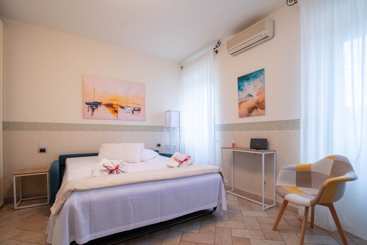 A well-lit living area is presented with a spacious bed dressed in fresh linens. Soft curtains frame a window, allowing natural light to brighten the room. A modern chair and a simple side table complement the decor, enhancing the overall comfort of the space.