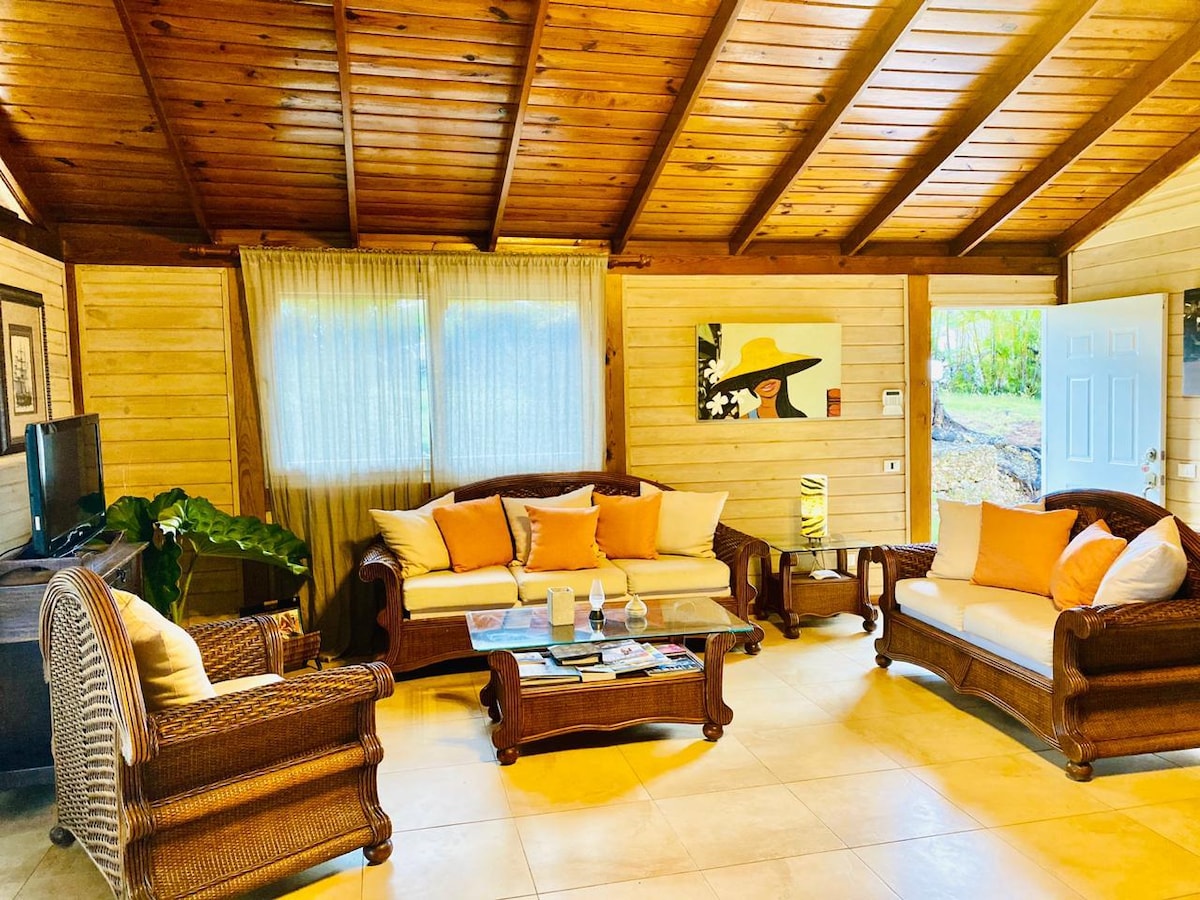 A cozy living area features a wooden ceiling with exposed beams, complemented by light-colored walls. Two wicker sofas adorned with orange cushions face a glass coffee table. A flat-screen TV is positioned beside a window with sheer curtains, allowing natural light to fill the space.
