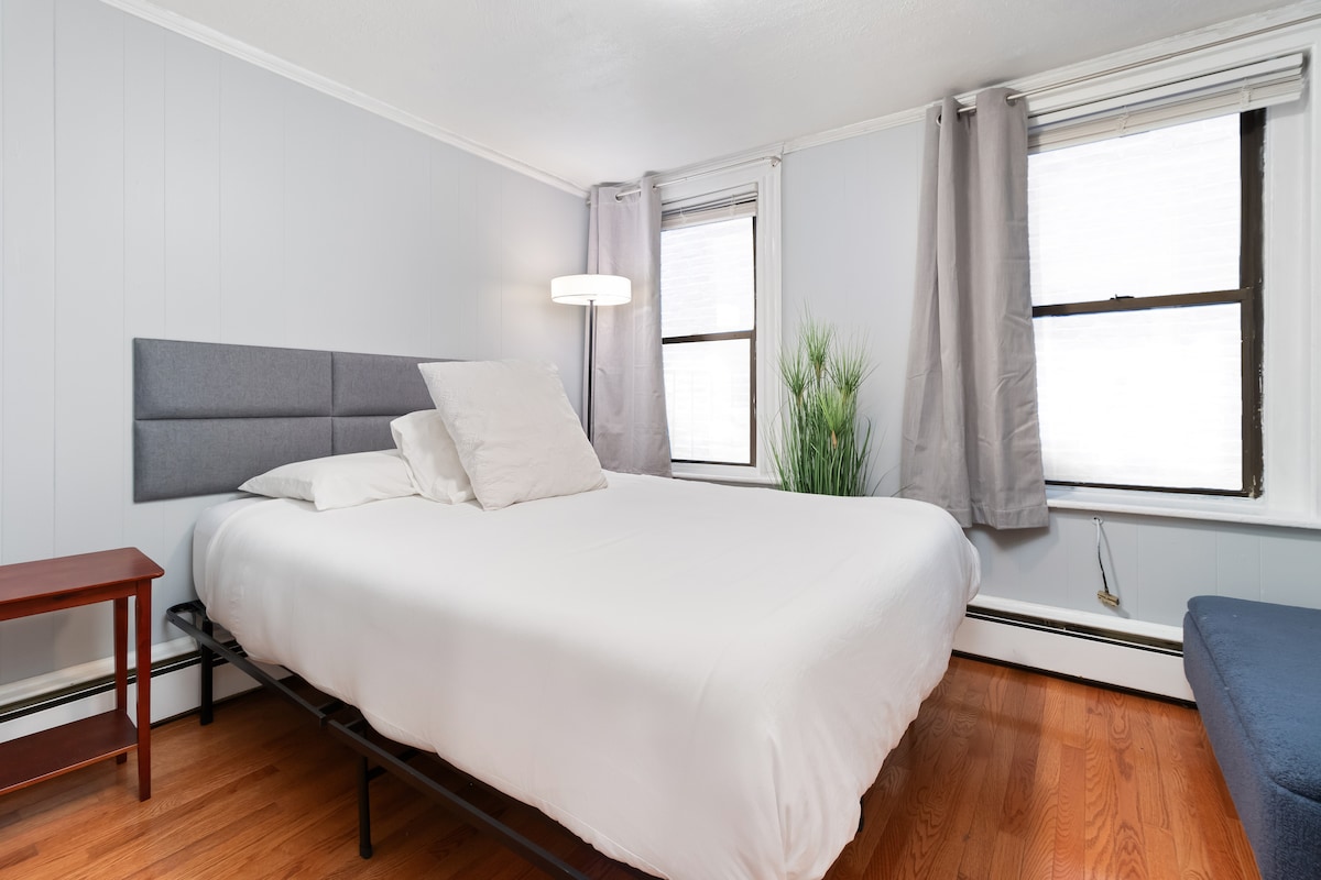 A bedroom features a spacious bed with light linens, complemented by accent pillows. Two windows are dressed with sheer curtains, allowing natural light to enter. A small bedside table is positioned next to the bed, and a modern floor lamp provides additional lighting.