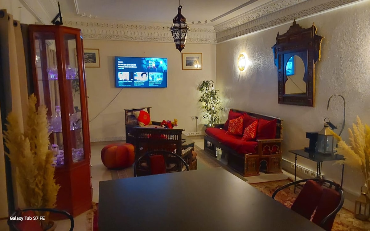 The living area features a traditional couch adorned with red and gold cushions, accompanied by dark wooden furniture. A flat-screen TV is mounted on the wall, with decorative elements and plants enhancing the environment. Soft lighting creates a warm ambiance throughout the space.