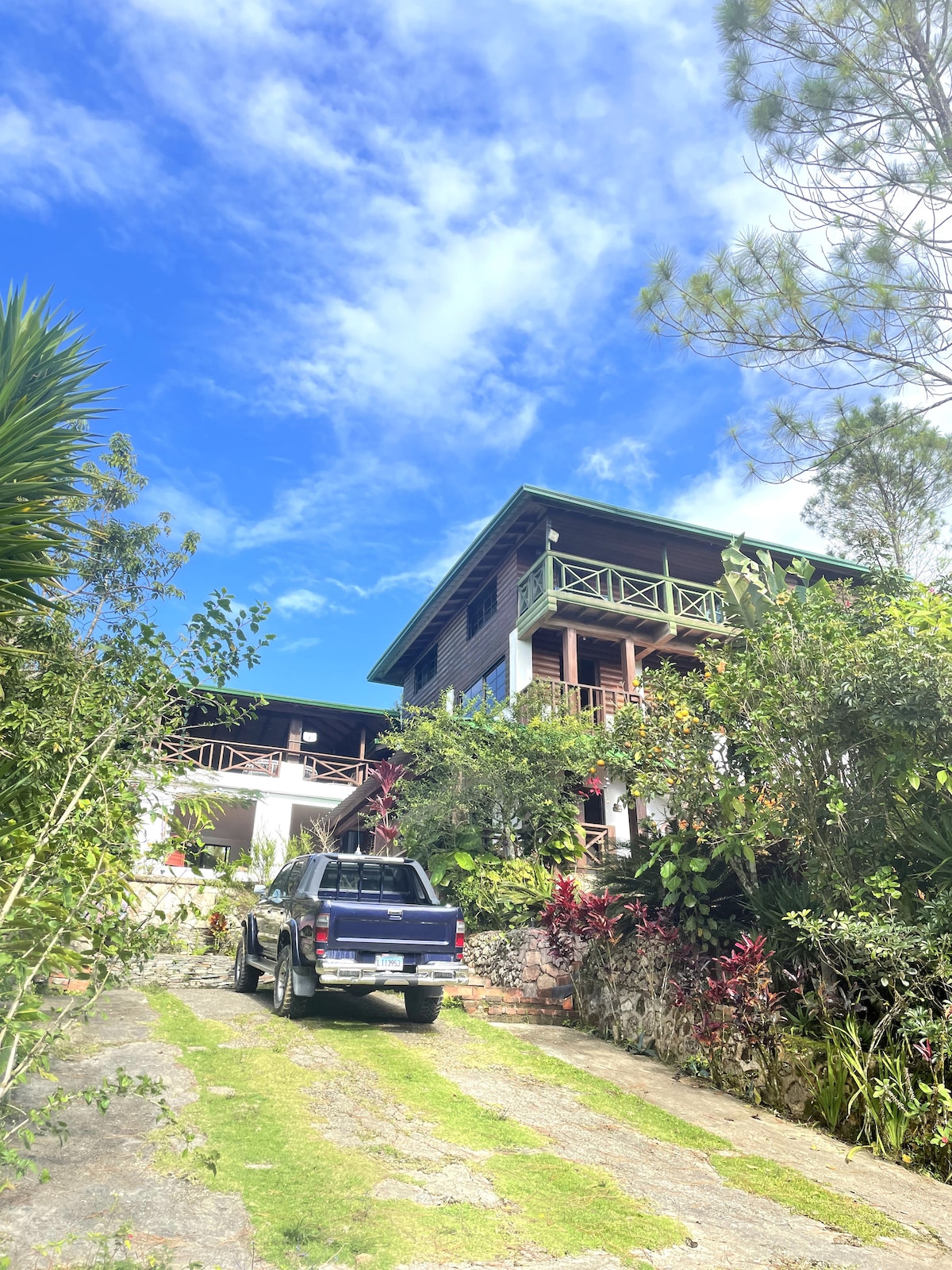 A three-level house is set against a clear blue sky, surrounded by lush green foliage and vibrant plants. A gravel driveway leads up to the home, which features wooden balconies and decorative railings. A black vehicle is parked in front, enhancing the rustic charm.