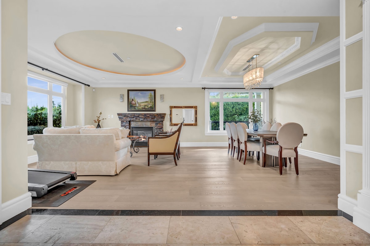 A spacious living area features a light-colored sofa set arranged around a decorative fireplace. A dining table with elegant chairs sits in the background, illuminated by a stylish chandelier. Large windows offer natural light and views of the outdoor greenery.