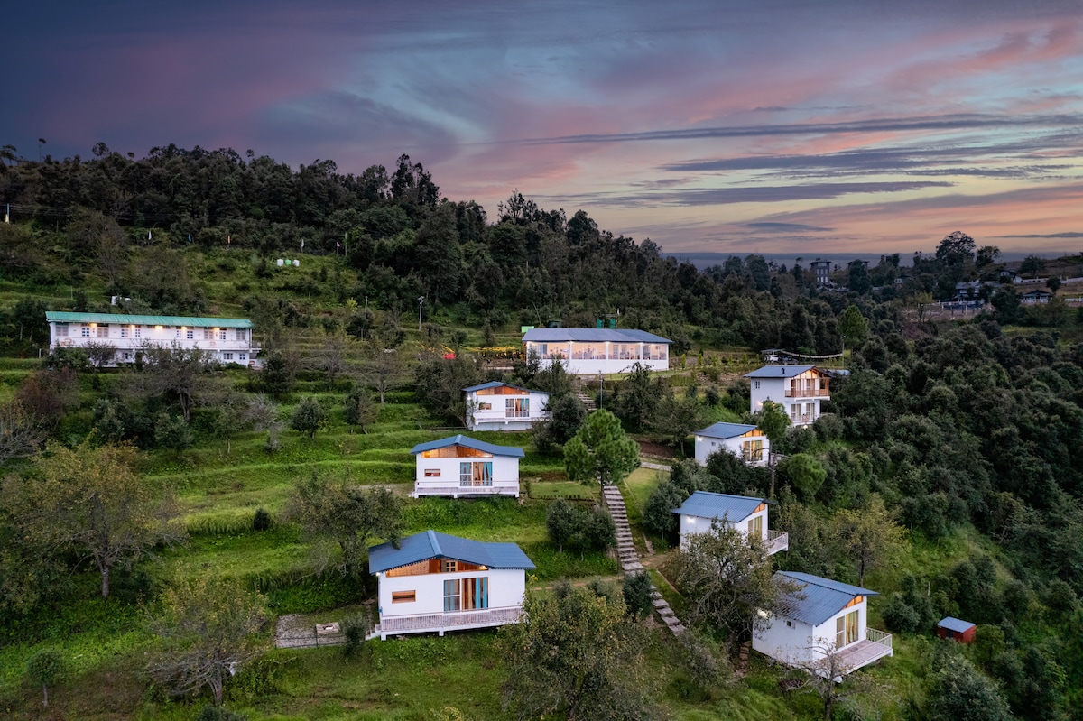 A scenic view of multiple cottages is captured on a hillside, surrounded by lush greenery and trees. The sky is painted with soft hues of sunset, adding warmth to the serene atmosphere. Pathways can be seen connecting the cottages, enhancing the inviting landscape.