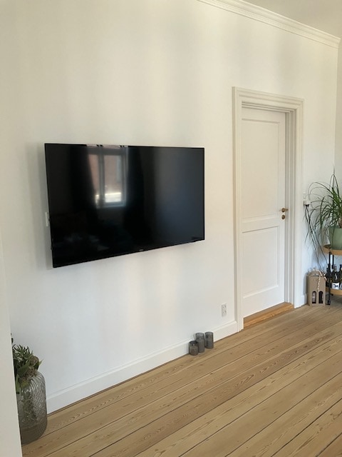 A wall-mounted television is located on a light-colored wall, opposite a closed white door. A potted plant and three candles are positioned on the floor beside the television, while wooden flooring adds a natural element to the space.