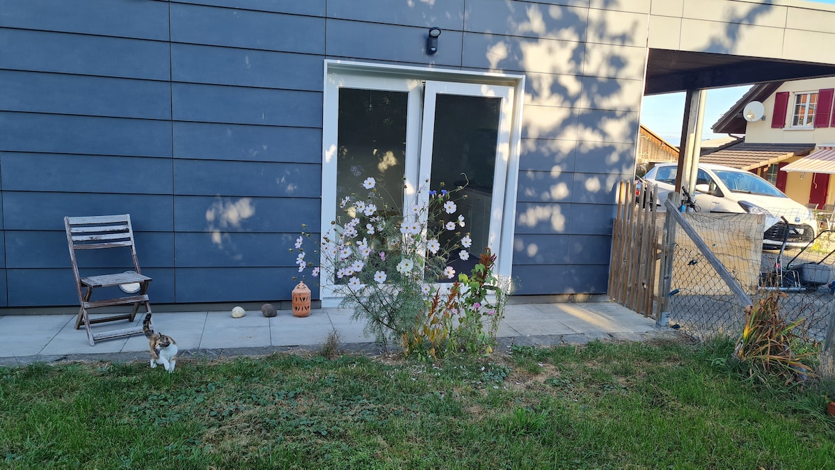 A private outdoor area is featured, showcasing a grassy space bordered by flowering plants. A small wooden chair is positioned beside the entrance, which is framed by large glass doors. A wooden fence separates the area from the neighboring property.