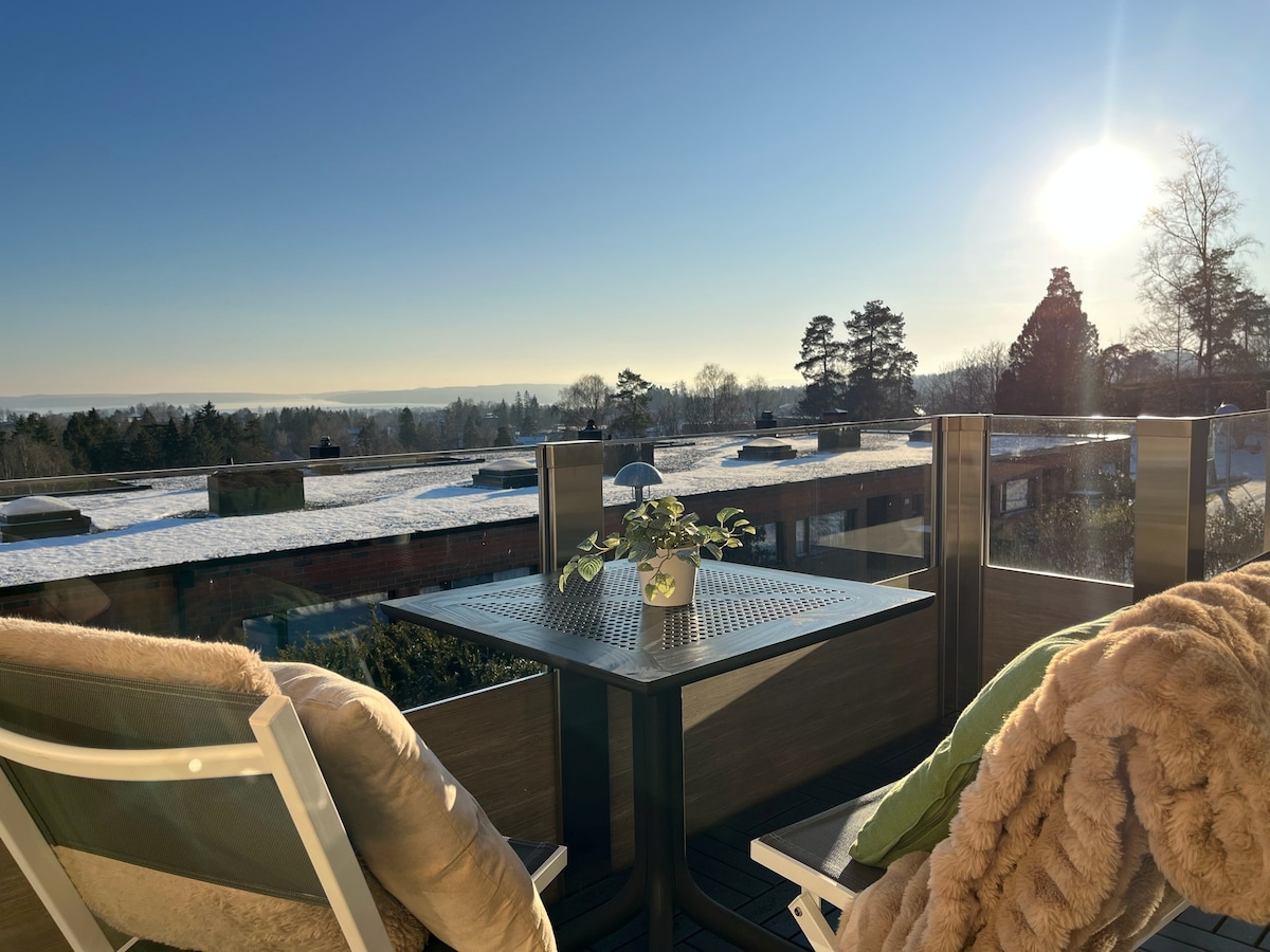 A balcony is featured with a small table and two chairs, accompanied by a cozy blanket and a plant centerpiece. A clear blue sky and sunlight create a bright atmosphere, while scenic views of trees and rooftops extend into the distance.