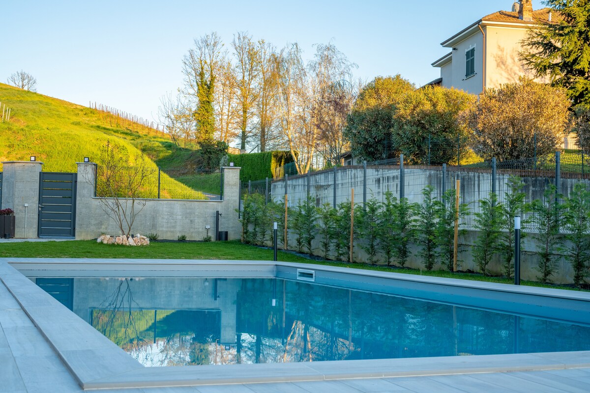 A clear swimming pool reflects the surrounding greenery and blue sky, set against a backdrop of gently sloping hills. The property is enclosed by a modern fence, with landscaped plantings enhancing the outdoor space.