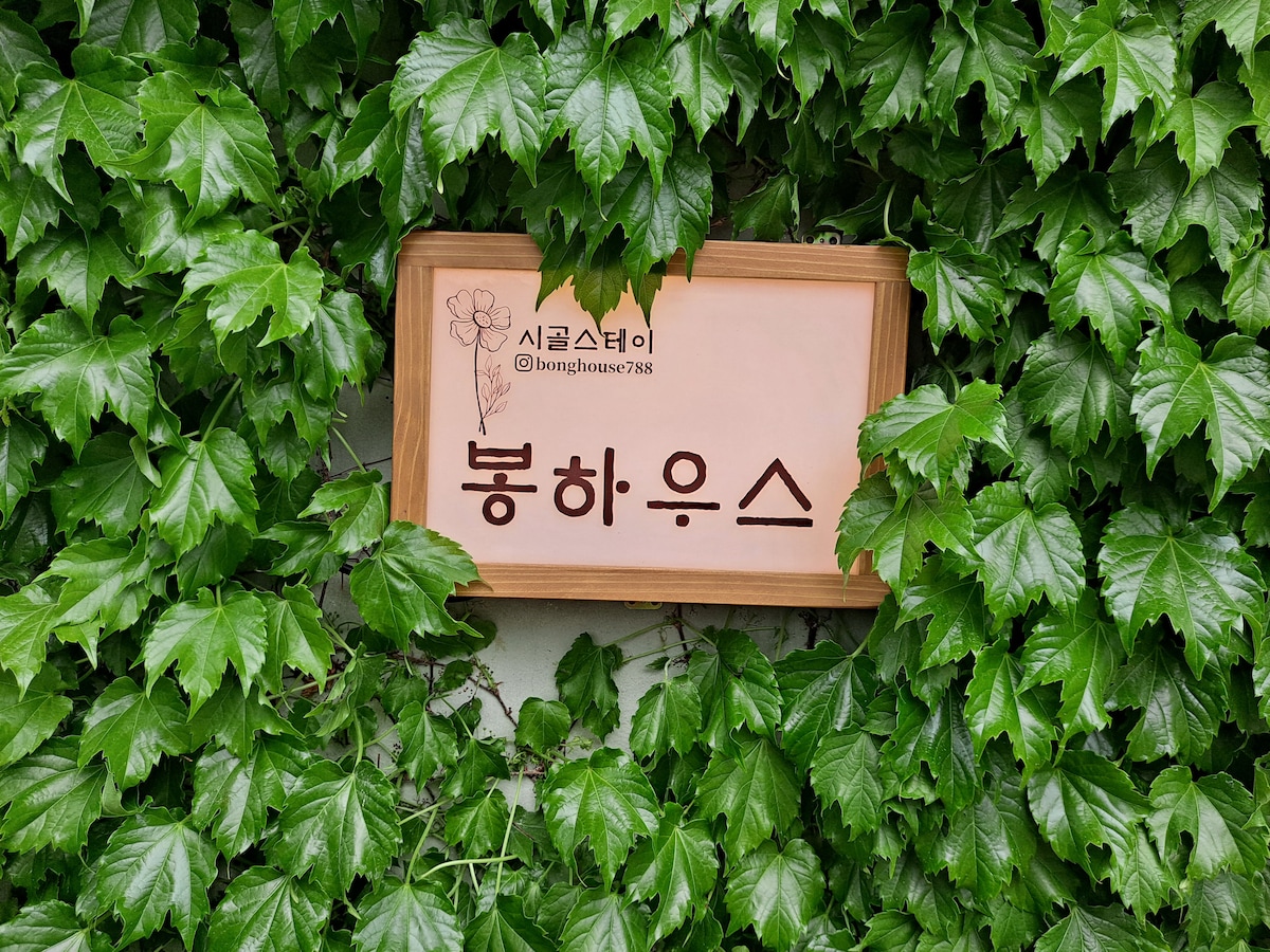 A wooden sign is nestled among vibrant green ivy, displaying the name '봉하우스' in a clear font. The lush leaves create a natural frame around the sign, enhancing the connection to the surrounding environment.