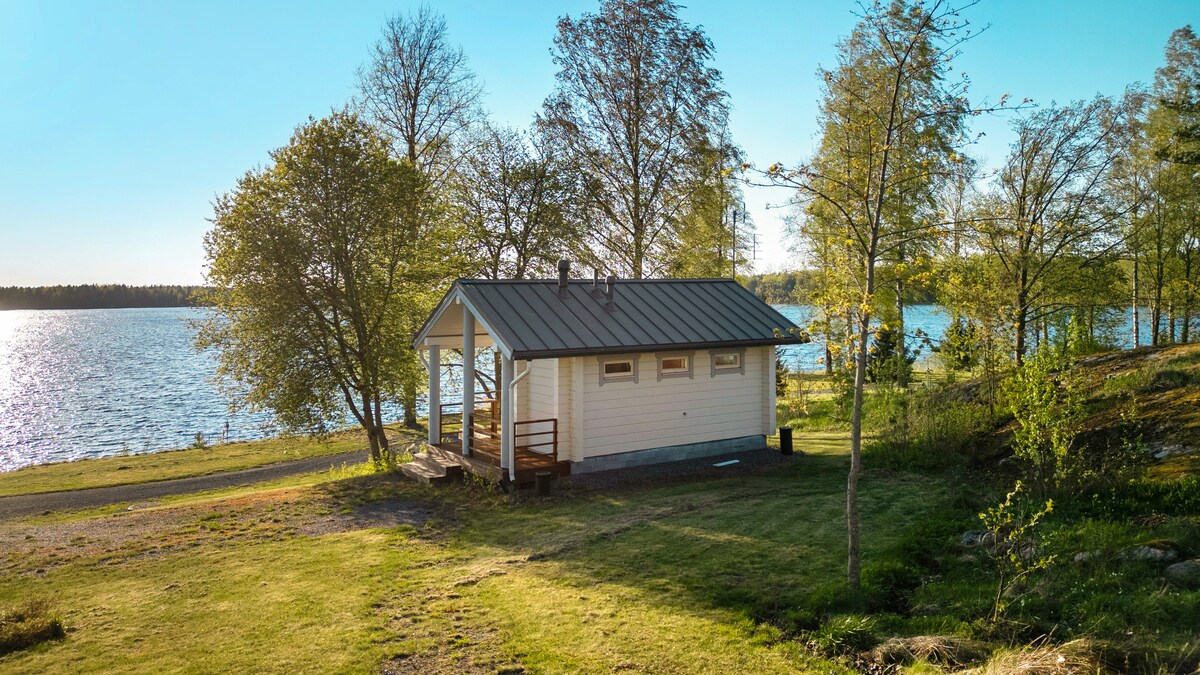 A compact cabin is situated near the water's edge, surrounded by lush green grass and trees. The structure features large windows that offer views of the lake. A covered porch extends from the front, providing a space to relax outdoors.