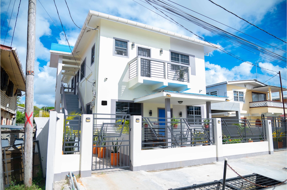 A two-story building is presented, featuring a modern exterior with a clean white finish and decorative blue accents. The entrance is flanked by steps and surrounded by decorative fencing. Sunlight illuminates the scene, highlighting the inviting outdoor space with potted plants.