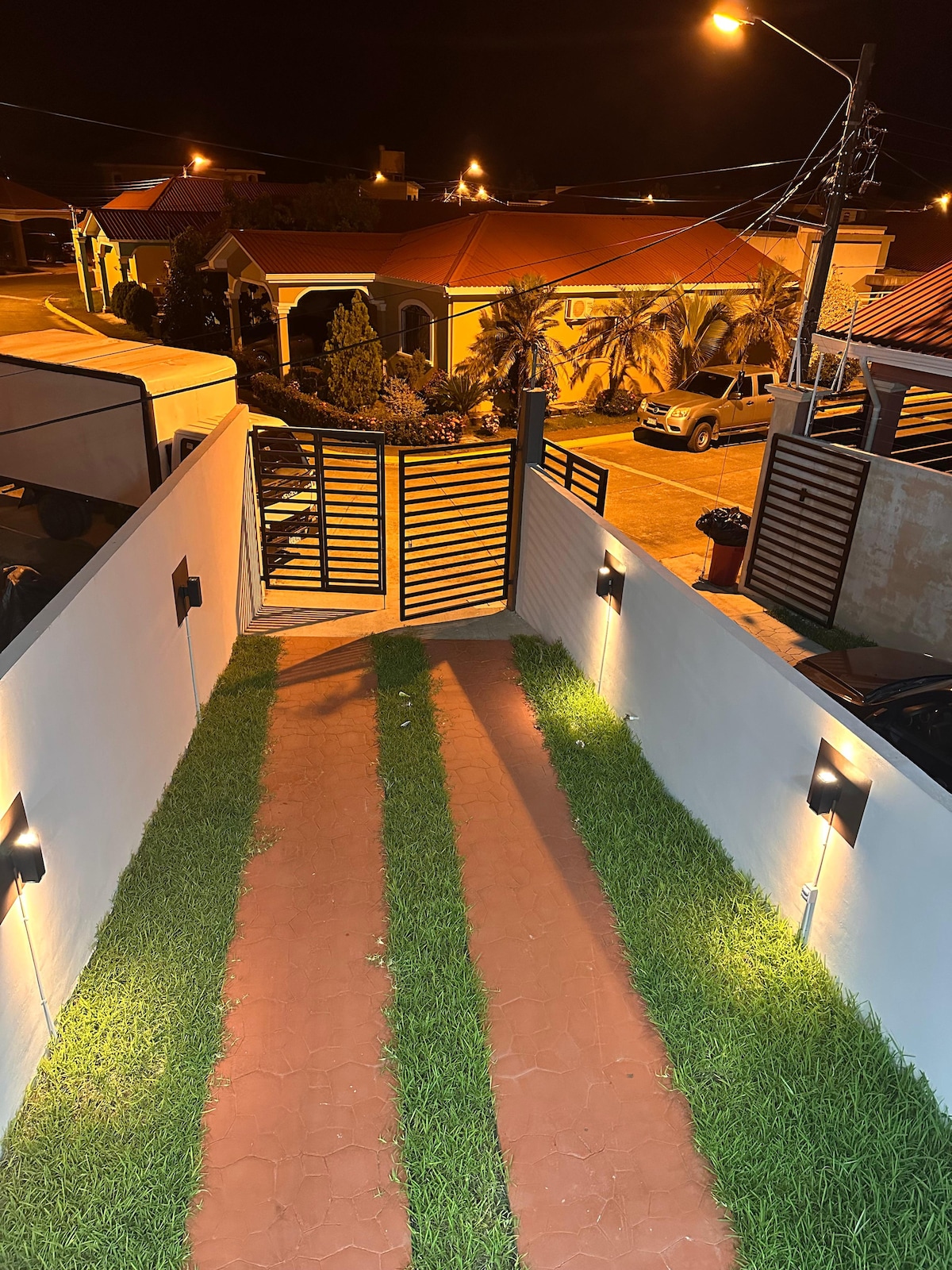A well-lit pathway features green grass flanking a red tile walkway, leading to an iron gate. Surrounding houses with sloped roofs are visible, illuminated by streetlights in the evening, providing a sense of safety and community.