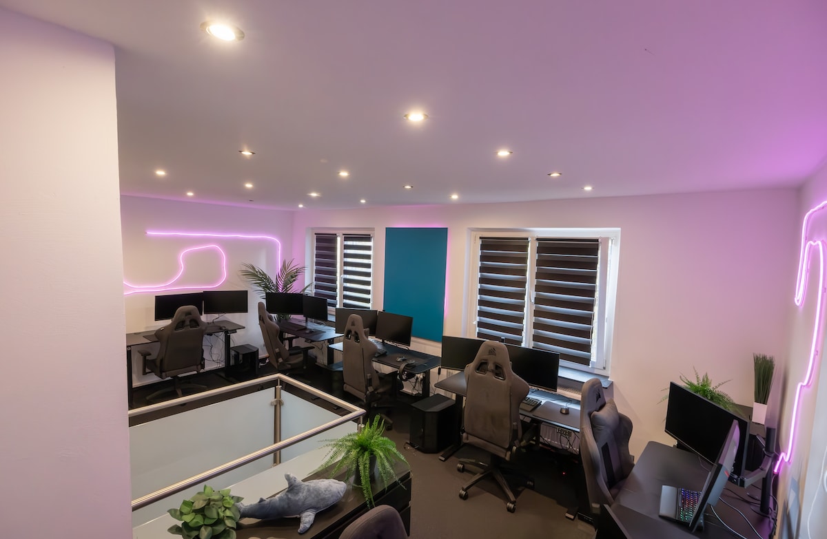 A modern workspace designed for gaming enthusiasts, featuring multiple ergonomic gaming chairs and workstations with large monitors. Soft pink lighting creates a motivating atmosphere. Two windows allow natural light, complemented by potted plants that add a touch of greenery to the environment.