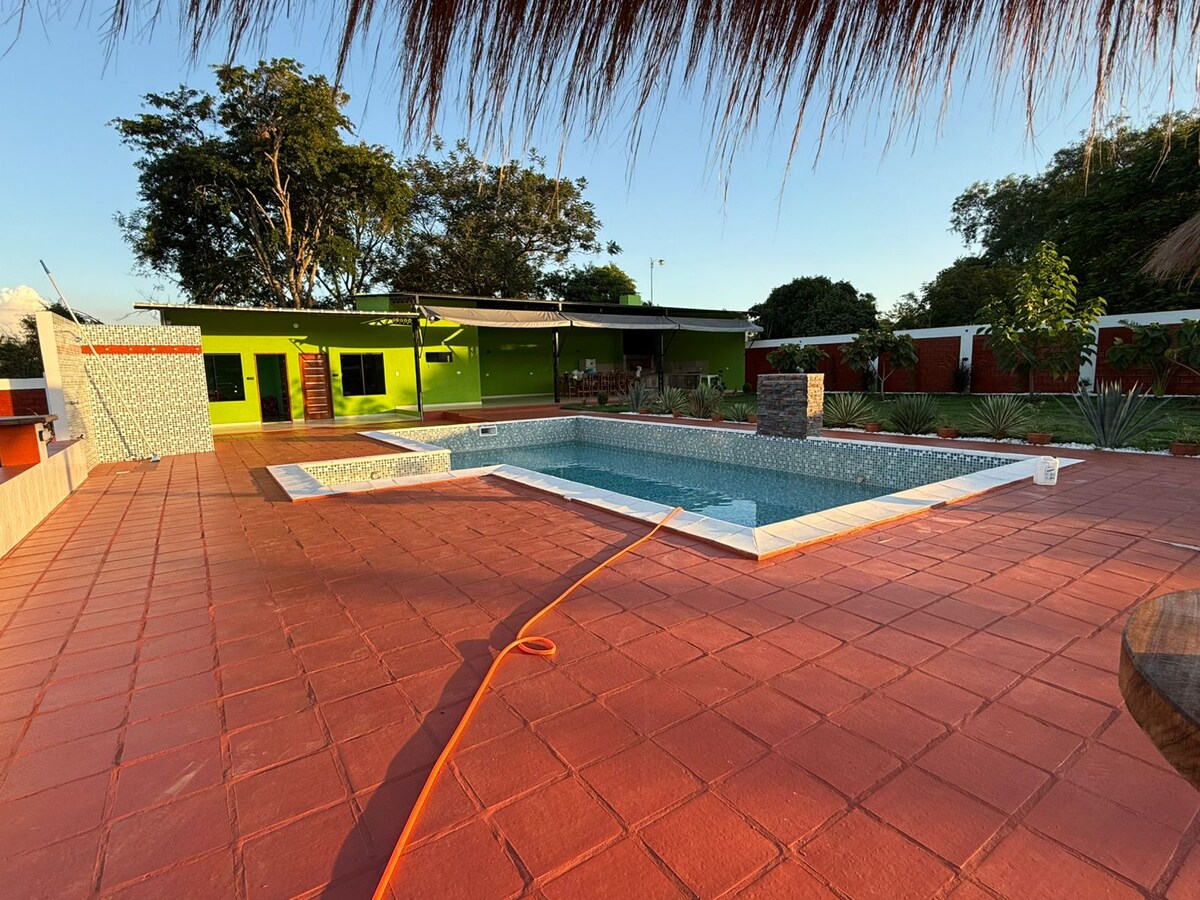 A spacious outdoor area features a rectangular pool surrounded by a vibrant red-tiled patio. Lush greenery and well-maintained gardens are visible in the background, with a modern building incorporating colorful walls and a shaded seating area nearby.